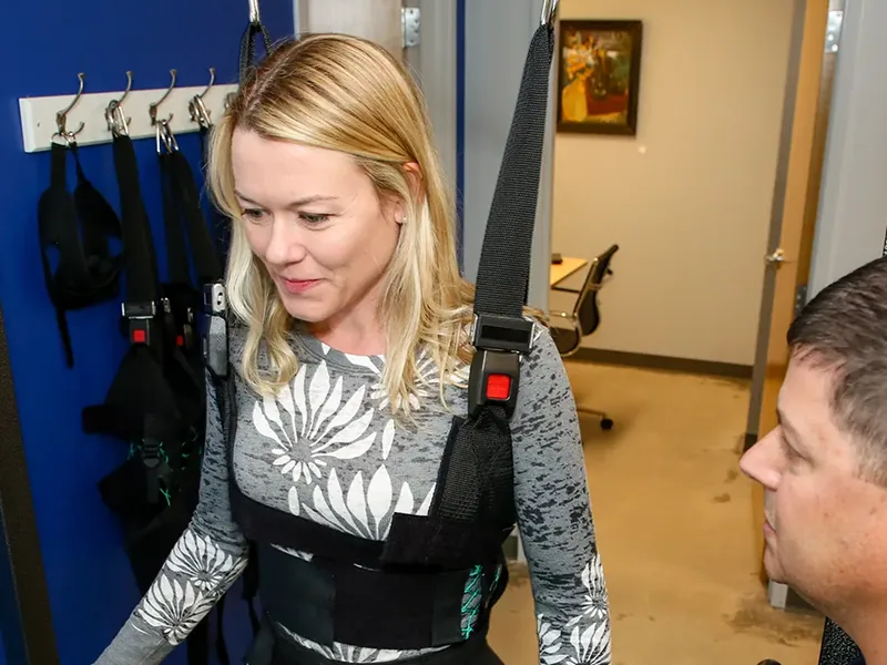 Patient supported by an overhead harness while a therapist stays alongside her for balance and walking practice
